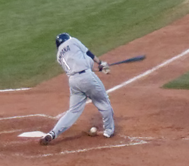Close up crop of photograph showing a baseball batter hitting a foul into the ground at his right foot, taken by an Olympus E-P3 in low light.